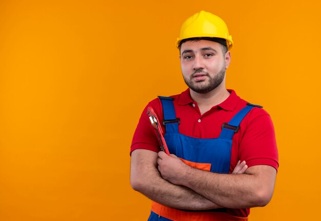 Homem jovem construtor satisfeito com uniforme de construção e capacete de segurança com as mãos cruzadas no peito, segurando a chave inglesa, parecendo confiante