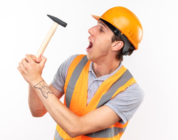 Foto grátis homem jovem construtor satisfeito de uniforme segurando e olhando para o martelo