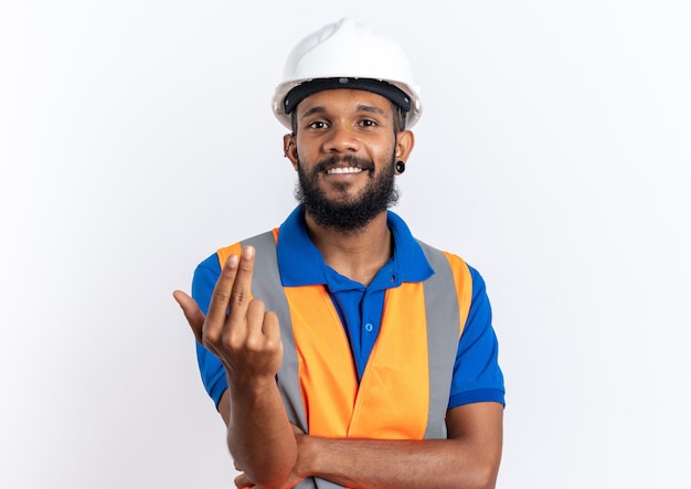 Homem jovem construtor sorridente, de uniforme com capacete de segurança, chamando alguém com a mão isolada na parede branca com espaço de cópia