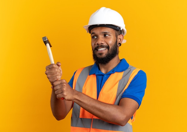 Homem jovem construtor sorridente de uniforme com capacete de segurança segurando e olhando para o martelo isolado na parede laranja com espaço de cópia