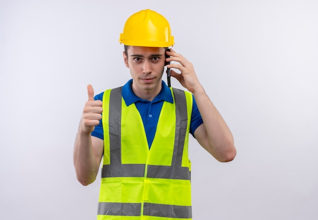 Foto grátis homem jovem construtor usando uniforme de construção e capacete de segurança, fazendo feliz sinal de positivo e falando ao telefone