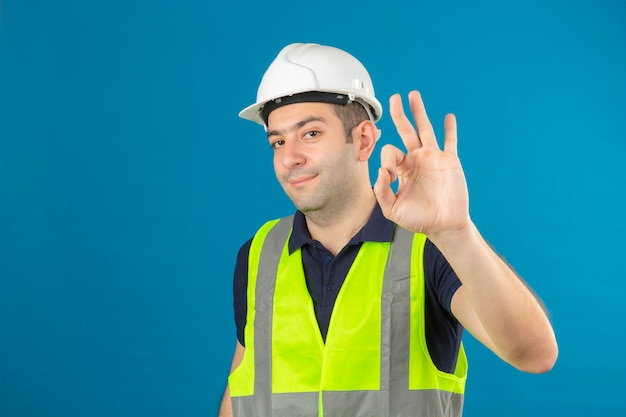 Homem jovem Construtor vestindo uniforme de construção e capacete de segurança no azul isolado positivo positivo fazendo sinal de ok com a mão e os dedos