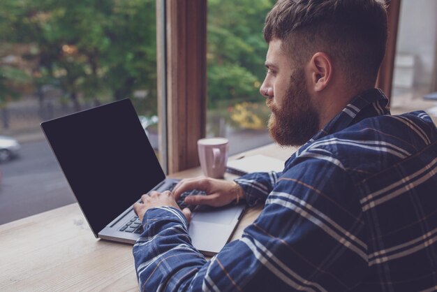 Homem jovem conversando via netbook durante o intervalo do trabalho em uma cafeteria, homem sentado em frente ao laptop aberto com a tela de cópia em branco para sua mensagem de texto ou conteúdo de publicidade