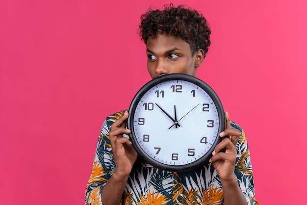 Homem jovem de pele escura, preocupado e pensativo, com cabelo encaracolado e camisa com estampa de folhas, segurando um relógio de parede que mostra as horas enquanto olha para o lado em um fundo rosa