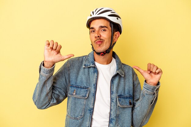 Homem jovem de raça mista usando uma bicicleta capacete isolada em fundo amarelo sente-se orgulhoso e autoconfiante, exemplo a seguir.
