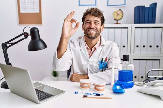 Homem jovem dentista hispânico trabalhando na clínica médica sorrindo positivo fazendo sinal de ok com expressão de sucesso de mão e dedos