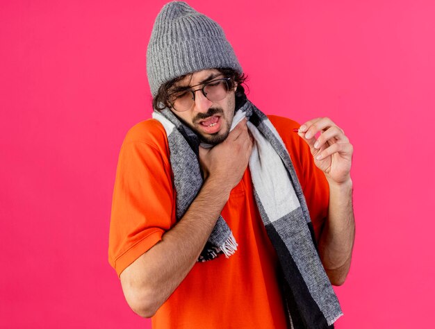 Homem jovem doente com óculos, chapéu de inverno e lenço segurando a garganta, mantendo as mãos no ar com os olhos fechados, isolado na parede rosa com espaço de cópia