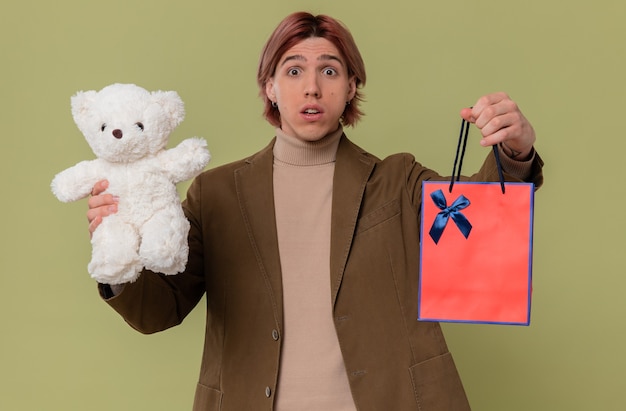Homem jovem e ansioso segurando um ursinho de pelúcia branco e uma sacola de presente