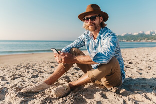 Homem jovem e atraente moderno sentado na praia à beira-mar nas férias de verão