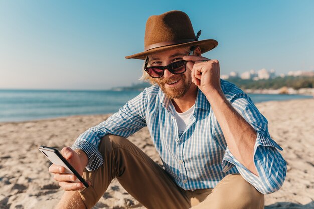 Homem jovem e atraente moderno sentado na praia à beira-mar nas férias de verão