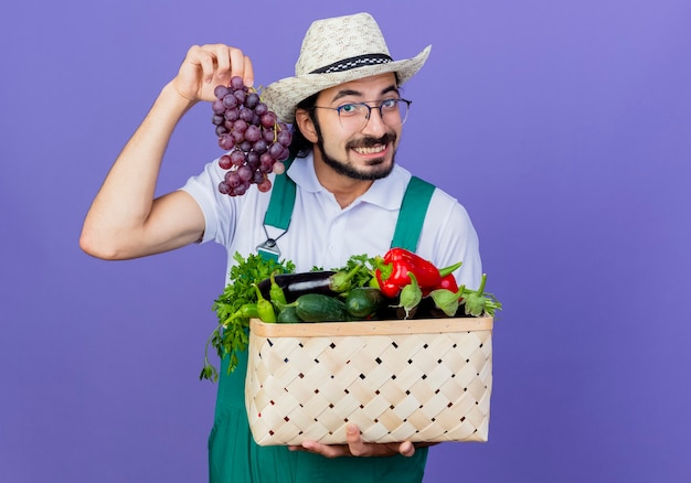 Homem jovem e barbudo jardineiro vestindo macacão e chapéu segurando uma caixa cheia de vegetais mostrando cacho de uva sorrindo em pé sobre a parede azul