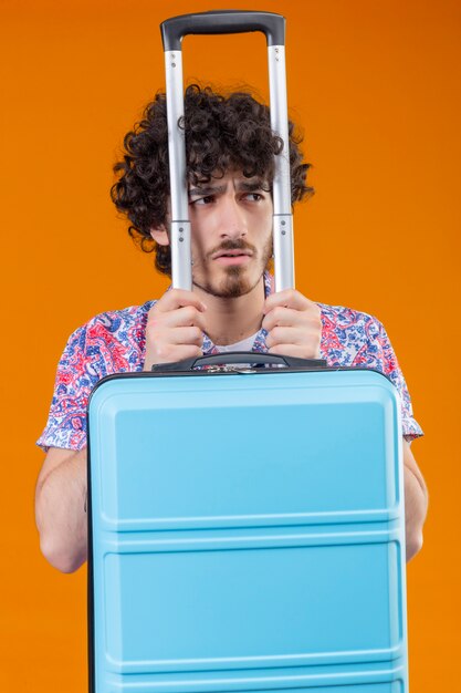 Homem jovem e bonito, desagradável, viajante encaracolado segurando uma mala, olhando para o lado direito de uma parede laranja isolada