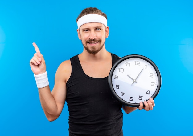 Foto grátis homem jovem e bonito e alegre, usando fita para a cabeça e pulseiras, segurando o relógio e apontando para cima, isolado no espaço azul