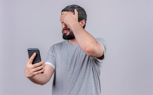 Homem jovem e bonito estressado segurando um telefone celular e colocando a mão na testa com os olhos fechados, isolado na parede branca