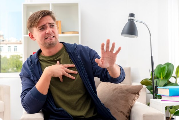 Homem jovem e bonito loiro irritado sentado na poltrona levantando a mão olhando para o lado