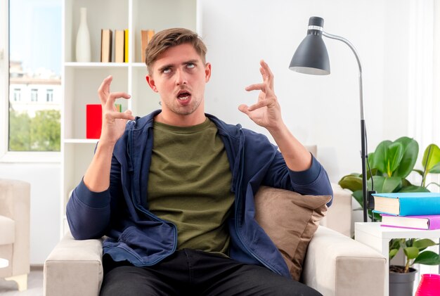 Homem jovem e bonito loiro irritado sentado na poltrona levantando as mãos e revirando os olhos dentro da sala