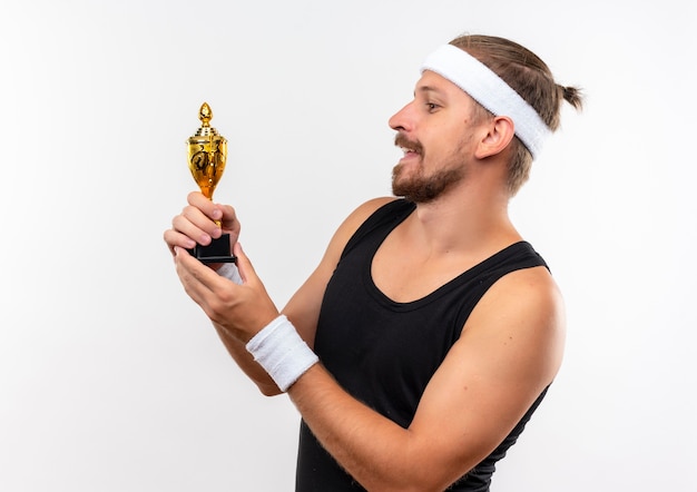 Foto grátis homem jovem e bonito satisfeito com a faixa na cabeça e pulseiras, segurando e olhando para a taça do vencedor, isolada no espaço em branco