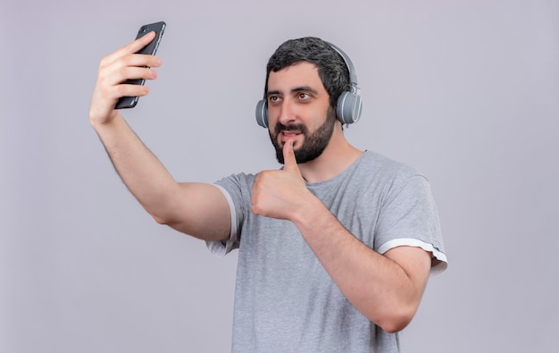 Homem jovem e bonito satisfeito usando fones de ouvido, mostrando o polegar para cima e tirando uma selfie isolada na parede branca