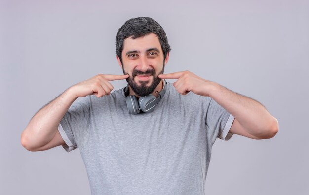 Homem jovem e bonito satisfeito usando fones de ouvido no pescoço e apontando para seu sorriso isolado na parede branca