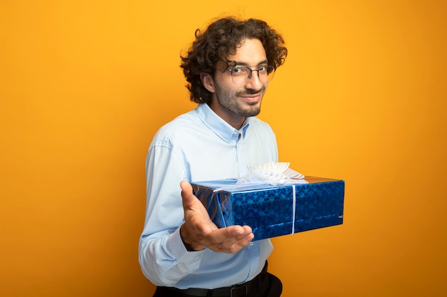 Homem jovem e bonito satisfeito, usando óculos, segurando um pacote de presente, olhando para a frente, isolado na parede laranja