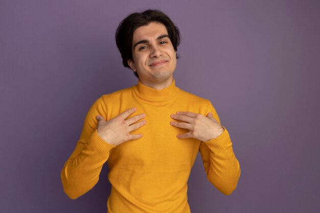 Homem jovem e bonito satisfeito vestindo uma blusa de gola alta amarela e colocando as mãos em si mesmo isolado na parede roxa