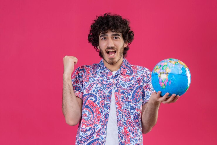 Foto grátis homem jovem e bonito viajante alegre segurando um globo levantando o punho em um espaço rosa isolado com espaço de cópia