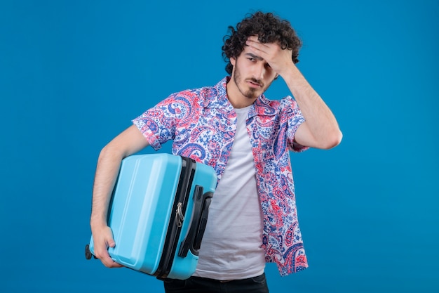 Foto grátis homem jovem e bonito viajante cansado segurando uma mala com a mão na testa em uma parede azul isolada com espaço de cópia