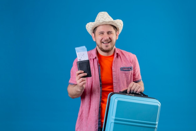 Homem jovem e bonito viajante com chapéu de verão segurando mala e passagens de avião, parecendo confiante e feliz, sorrindo alegremente em pé sobre a parede azul Foto gratuita