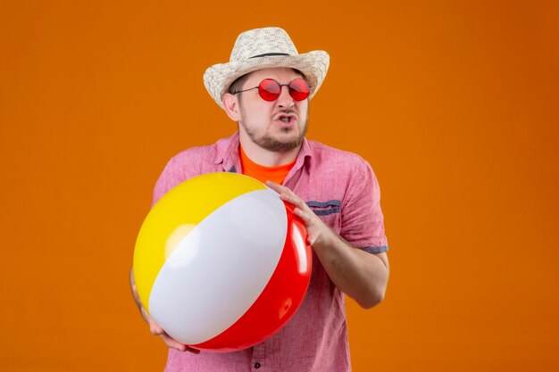 Homem jovem e bonito viajante com chapéu de verão segurando uma bola inflável e se sentindo irritado