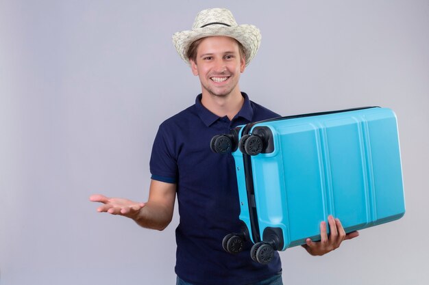 Homem jovem e bonito viajante com chapéu de verão segurando uma mala e sorrindo alegremente para a câmera em pé com o braço levantado sobre um fundo branco