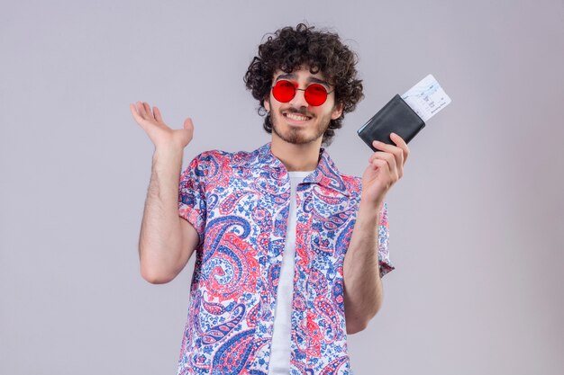 Homem jovem e bonito viajante encaracolado sorridente, usando óculos escuros, segurando uma carteira e bilhetes de avião e mostrando a mão vazia na parede branca isolada