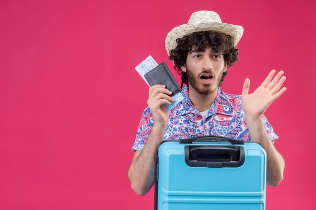 Homem jovem e bonito viajante encaracolado surpreso, usando um chapéu segurando passagens de avião e carteira com mala na parede rosa isolada com espaço de cópia