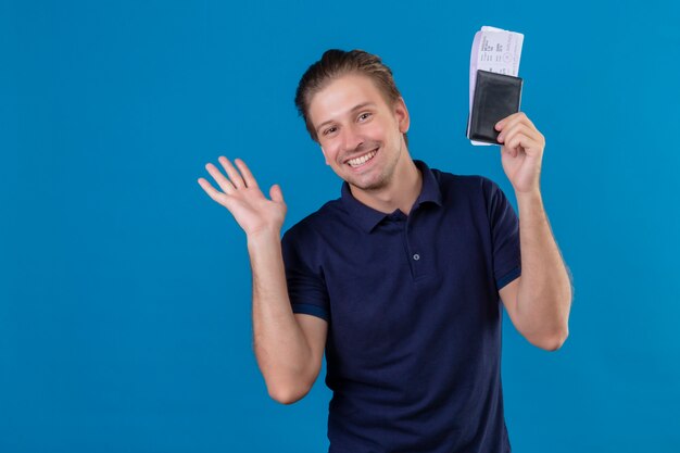 Homem jovem e bonito viajante satisfeito segurando passagens aéreas e sorrindo alegremente, olhando para a câmera acenando com a mão em pé sobre um fundo azul