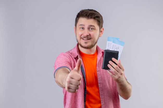 Homem jovem e bonito viajante segurando passagens aéreas, sorrindo de forma positiva e feliz, mostrando os polegares em pé sobre uma parede branca