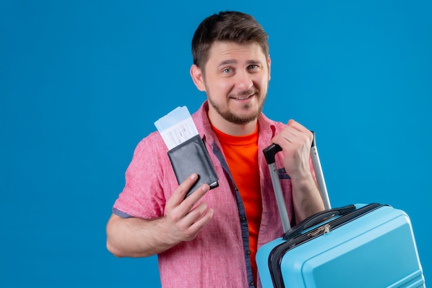 Homem jovem e bonito viajante segurando passagens de avião e mala sorrindo positivamente