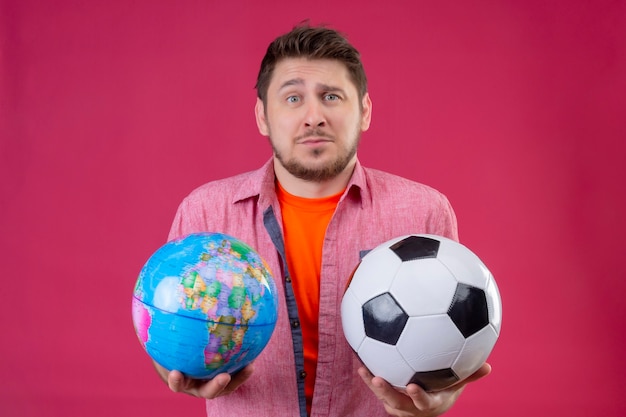 Homem jovem e bonito viajante segurando uma bola de futebol e um globo, parecendo confuso e incerto