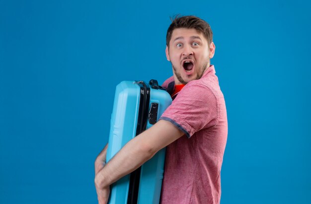 Homem jovem e bonito viajante segurando uma mala azul chocado com a boca aberta