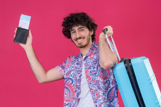 Homem jovem e bonito viajante sorridente segurando a carteira, as passagens de avião e a mala na parede rosa isolada