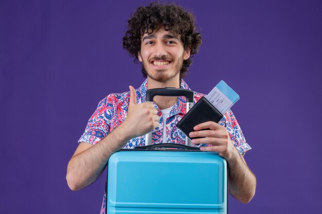 Homem jovem e bonito viajante sorridente segurando passagens de avião e carteira com a mala aparecendo com o polegar na parede roxa isolada