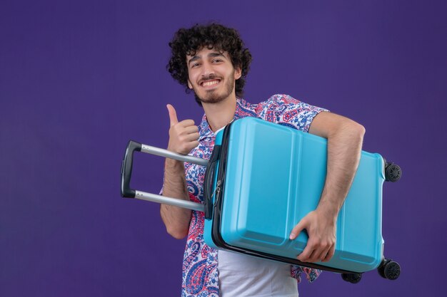 Foto grátis homem jovem e bonito viajante sorridente segurando uma mala e mostrando o polegar na parede roxa isolada com espaço de cópia