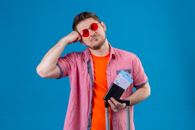 Foto grátis homem jovem e bonito viajante usando óculos escuros segurando passagens de avião com uma expressão triste no rosto em pé sobre a parede azul