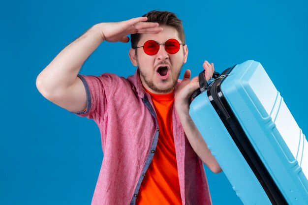Homem jovem e bonito viajante usando óculos escuros segurando uma mala, parecendo surpreso e surpreso ao lado da parede azul