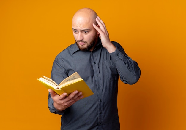 Homem jovem e careca de call center impressionado segurando e olhando para um livro com a mão na cabeça isolada em laranja com espaço de cópia