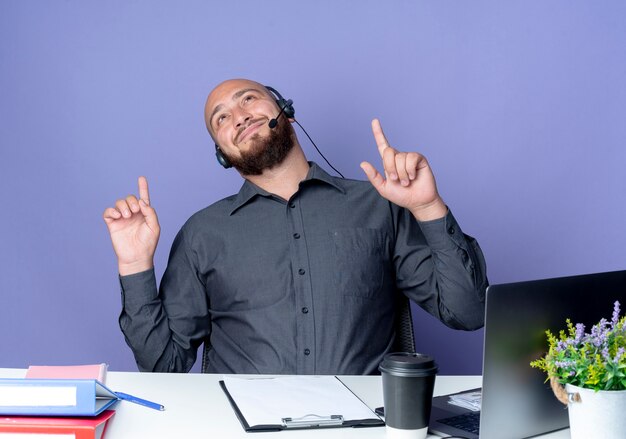 Homem jovem e careca satisfeito do call center usando fone de ouvido, sentado na mesa com ferramentas de trabalho, olhando e apontando para cima, isolado no fundo roxo