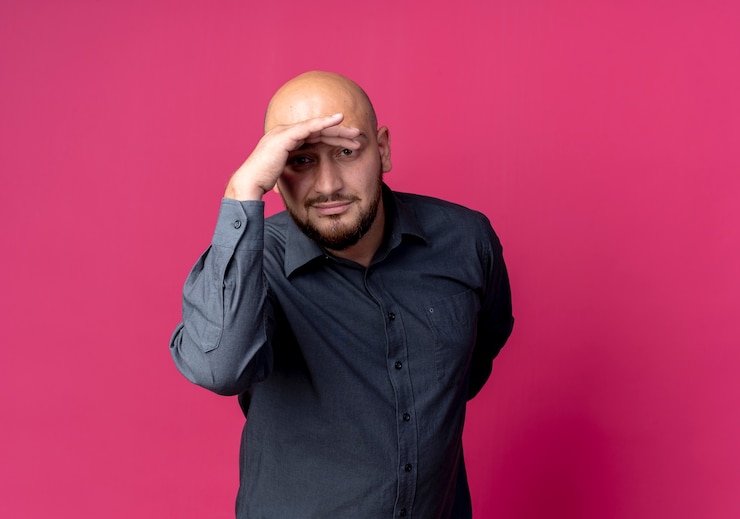Foto grátis homem jovem e careca satisfeito pelo call center, olhando para a distância e mantendo a mão atrás das costas, isolado em um fundo carmesim com espaço de cópia