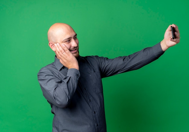 Homem jovem e careca sorridente, colocando a mão no rosto e tirando uma selfie isolada na parede verde