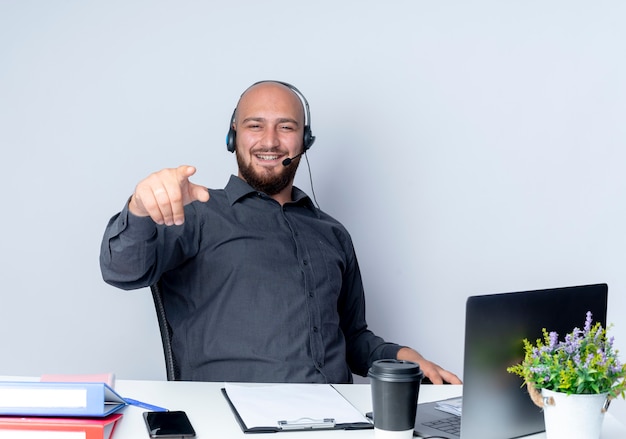 Homem jovem e careca sorridente de call center usando fone de ouvido sentado na mesa com ferramentas de trabalho apontando para frente, isoladas na parede branca