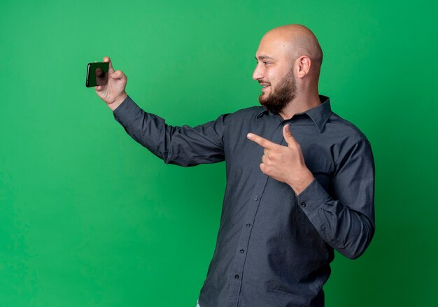 Homem jovem e careca sorridente, tirando uma selfie e apontando para a frente, isolado na parede verde