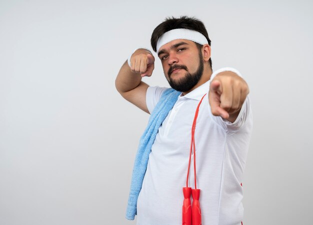 Homem jovem e confiante e esportivo usando bandana e pulseira com toalha e pular corda no ombro, mostrando seu gesto isolado na parede branca com espaço de cópia