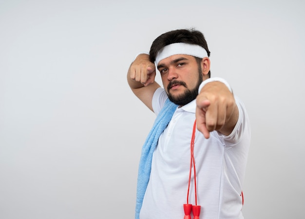 Homem jovem e confiante e esportivo usando bandana e pulseira com toalha e pular corda no ombro, mostrando seu gesto isolado na parede branca com espaço de cópia
